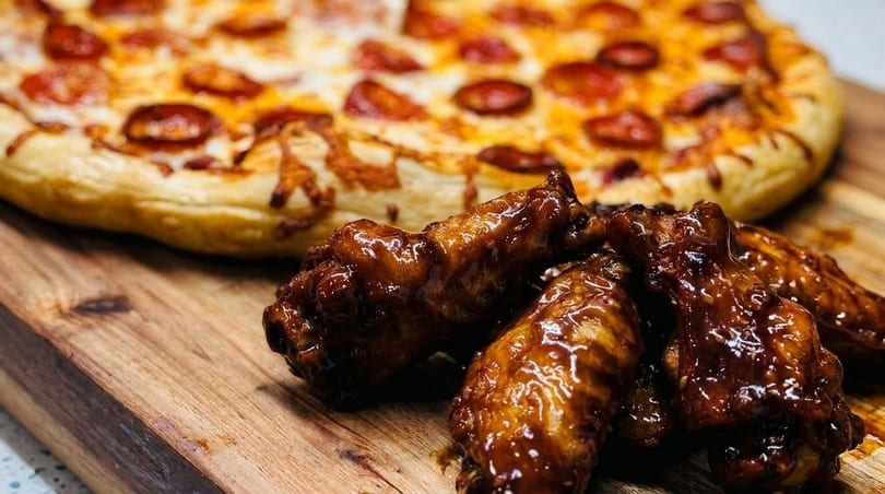 Glossy BBQ chicken wings and a pepperoni pizza on a wooden cutting board.